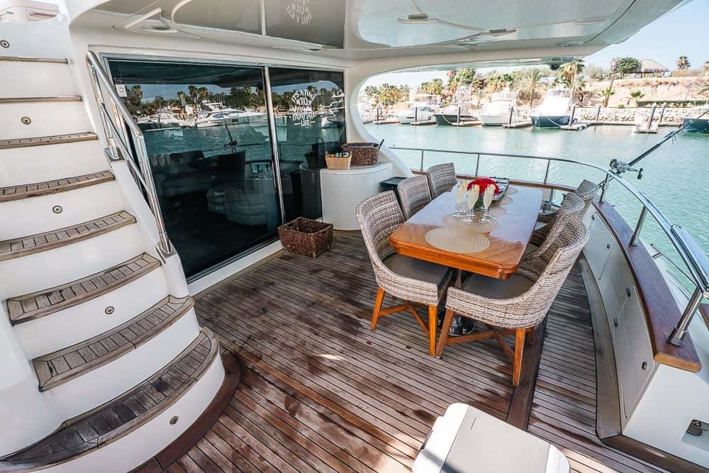 Aft deck with a teak floor, a dining table with woven chairs and the stairs up to the flybridge, the marina behind