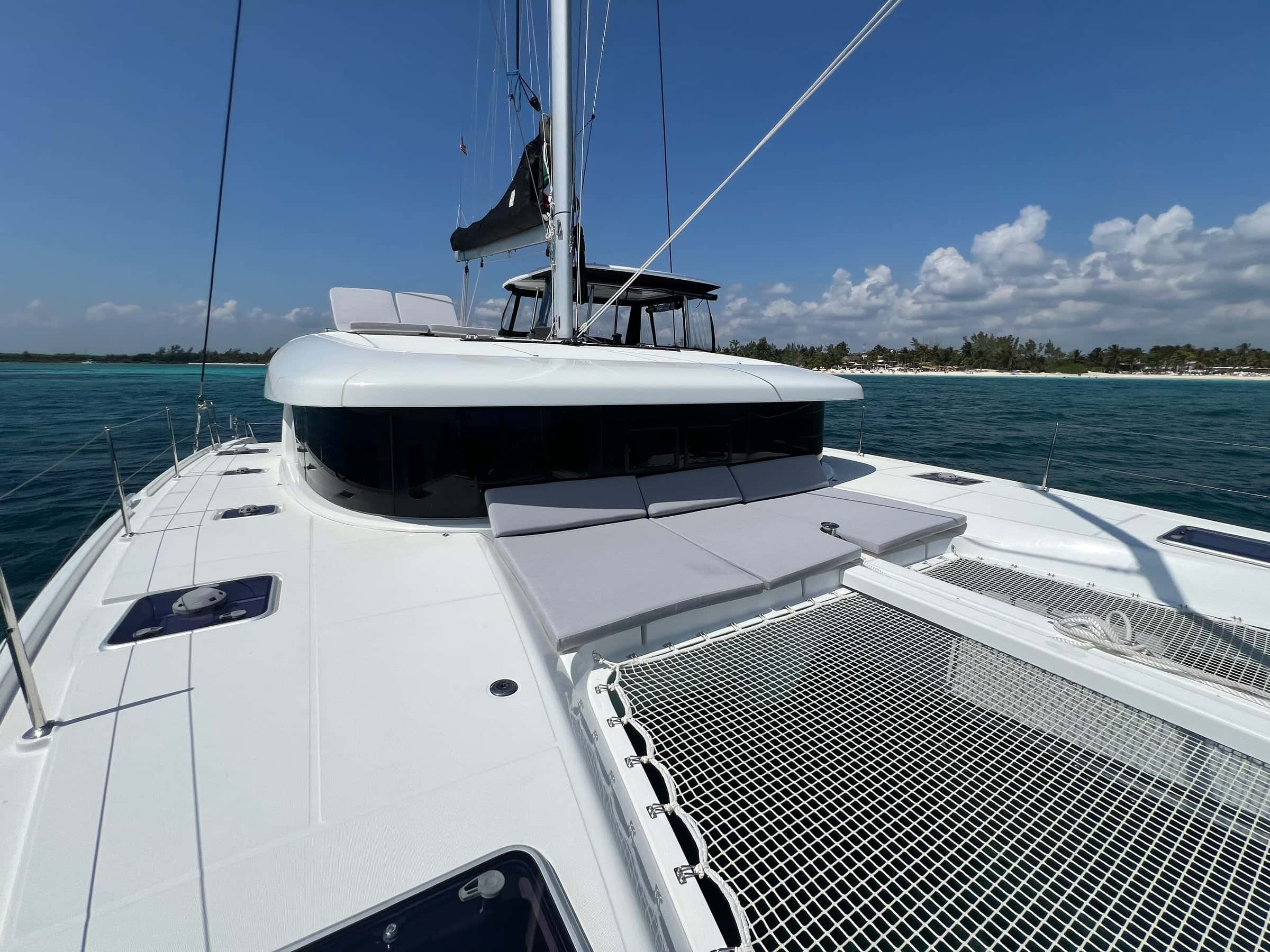 Grey sun pads at the front of the coachroof above the bow net, a palm-lined beach across the water