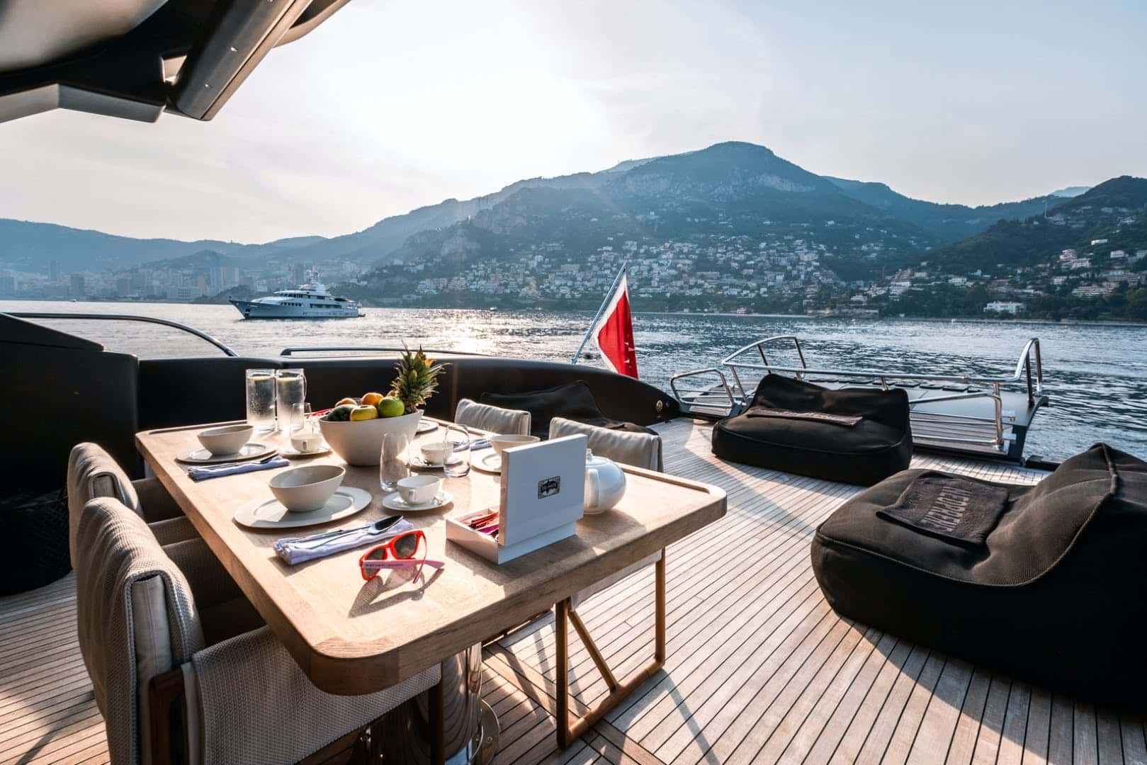 Breakfast table with a fruit bowl on a teak aft deck, bean bags beside it and a hillside town behind