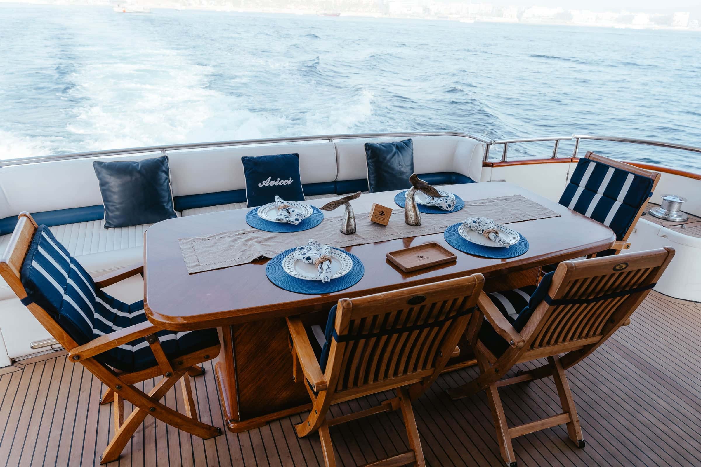 Aft deck teak table set with blue plates and napkins, folding teak chairs and a cushioned bench, the wake beyond