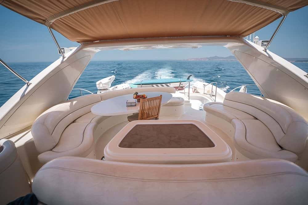 Flybridge lounge with a U-shaped cream settee around a table under the hardtop, the wake behind