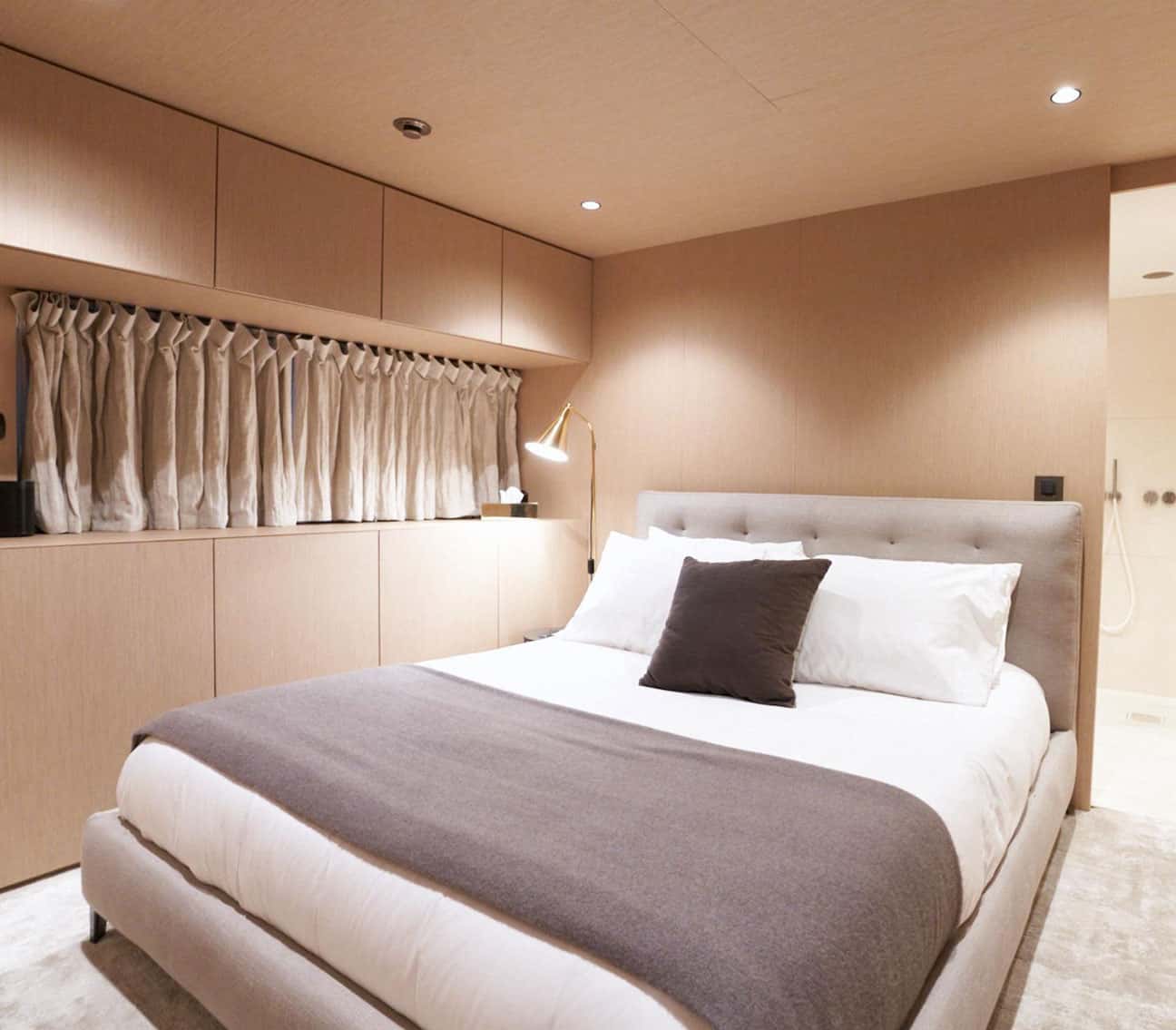 Double cabin with a grey upholstered bed, linen curtains, wood cabinets and a shower beyond