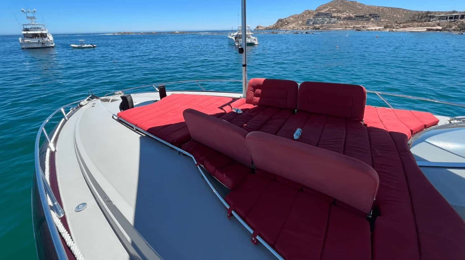 Bow lounge covered in red cushions with backrests, looking out over turquoise water and a rocky headland