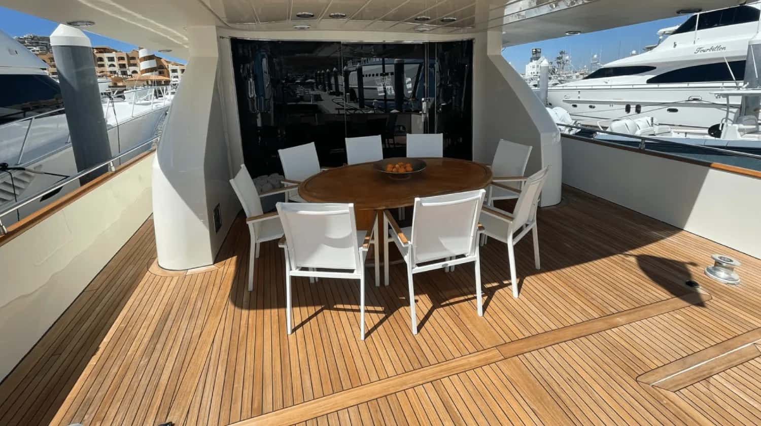 Covered aft deck in the marina with a round teak table, eight white chairs and a boat beside it