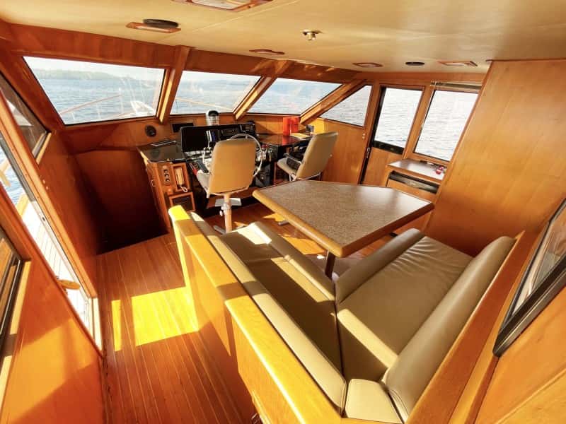 Sunlit pilothouse with two helm chairs, a table and a settee, and water views