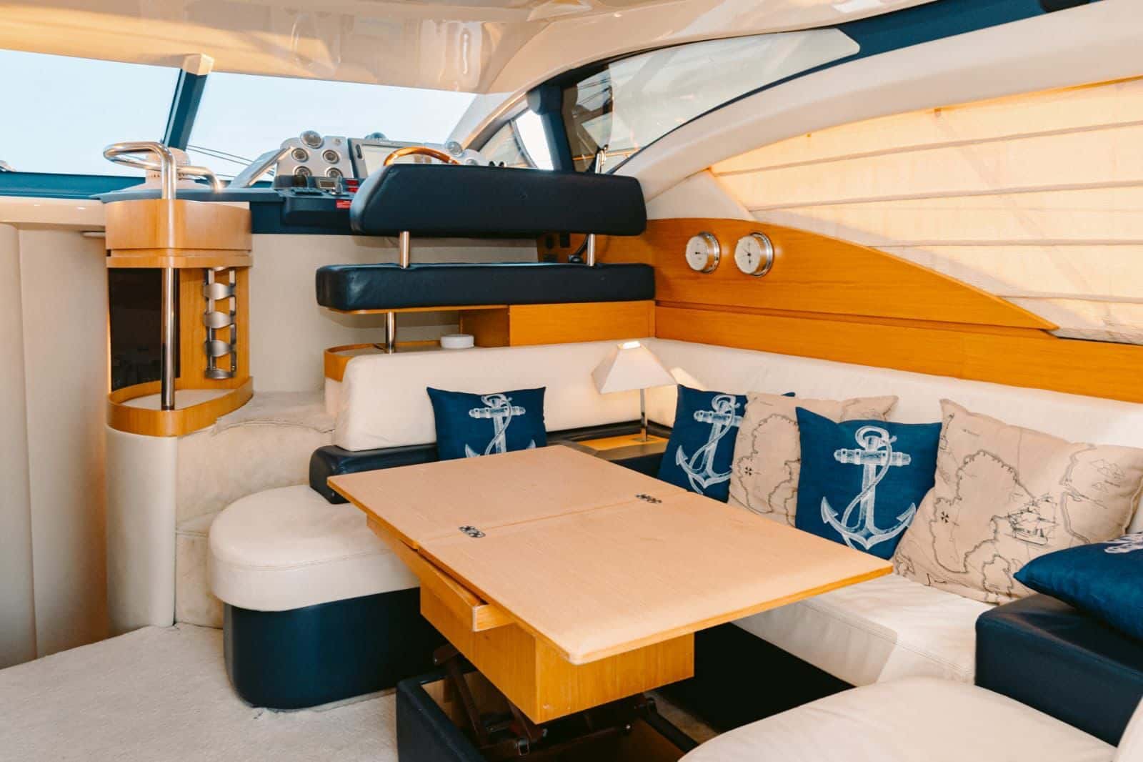 Saloon with U-shaped cream settee, folding wooden table, anchor cushions and helm behind