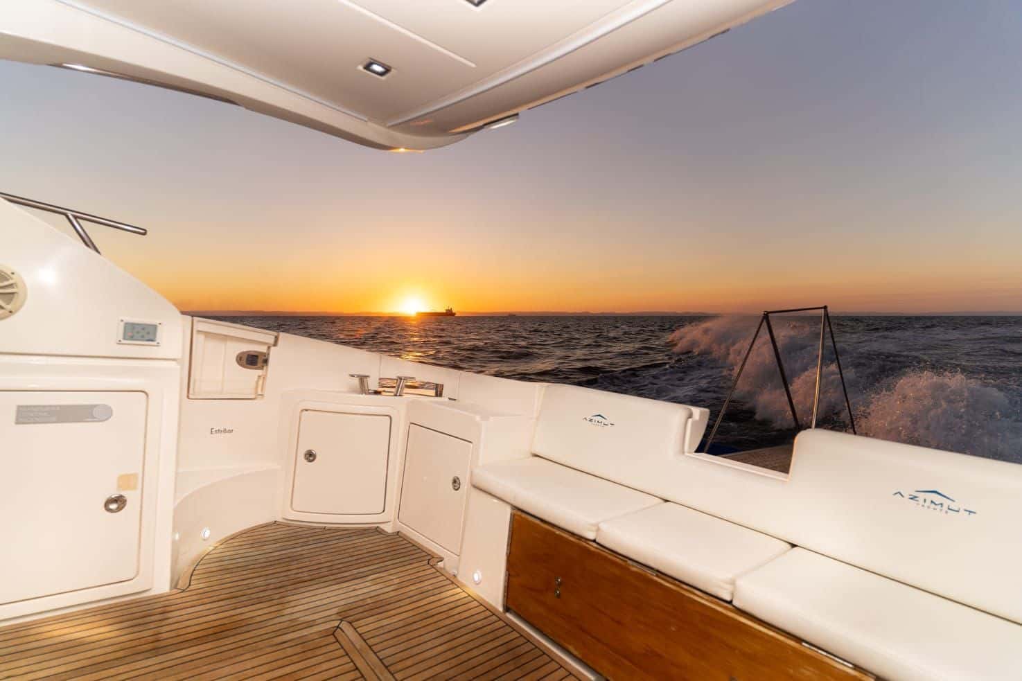 Stern cockpit with white cushioned bench seating and teak deck at sunset, wake behind