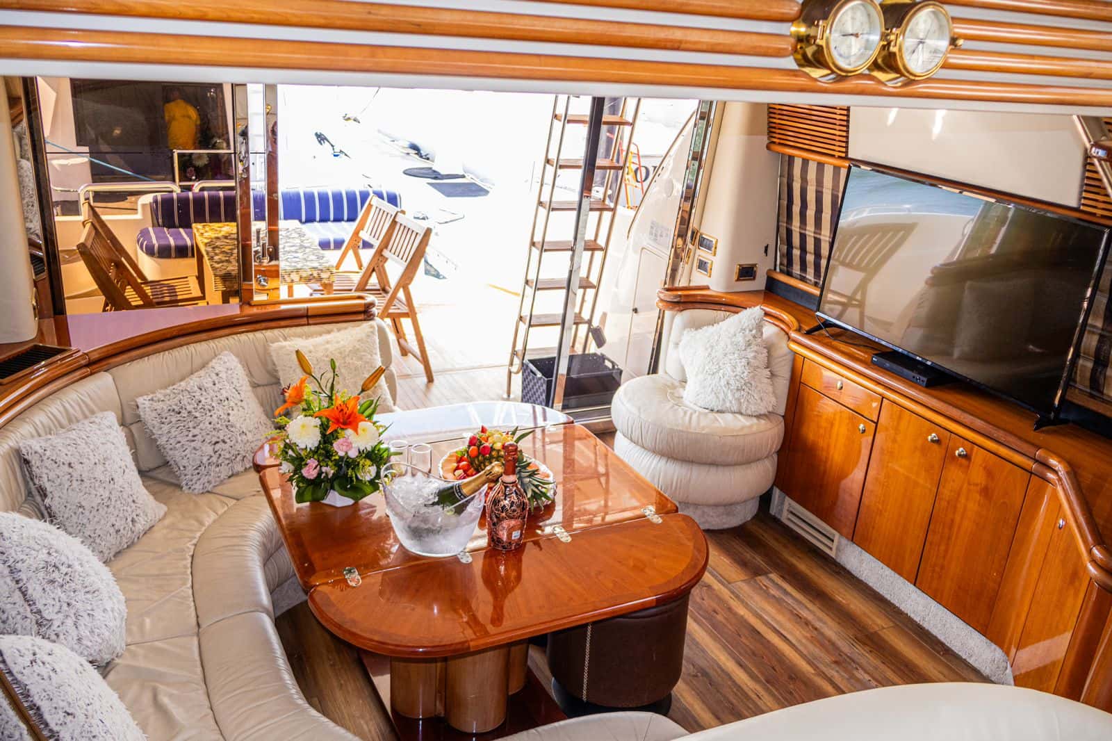 Saloon with a curved cream sofa, wooden table with champagne and fruit, a flat-screen TV and stern doors