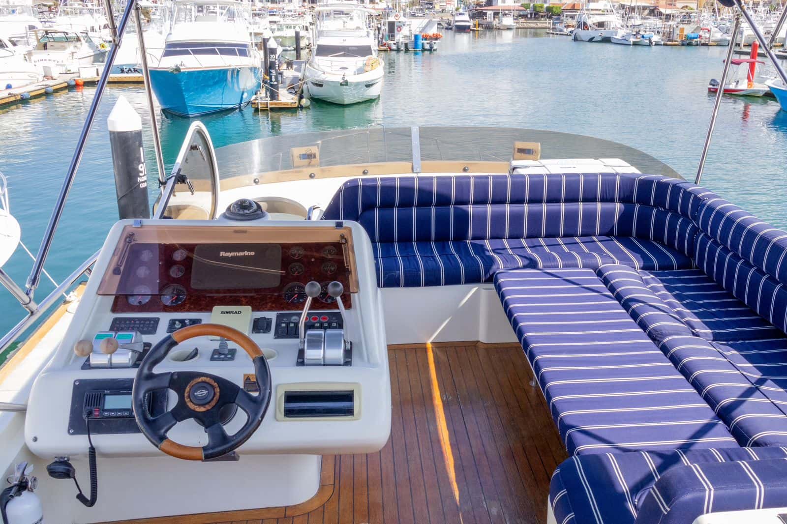 Flybridge with the helm console and a large navy striped U-shaped sofa under a Bimini, marina beyond