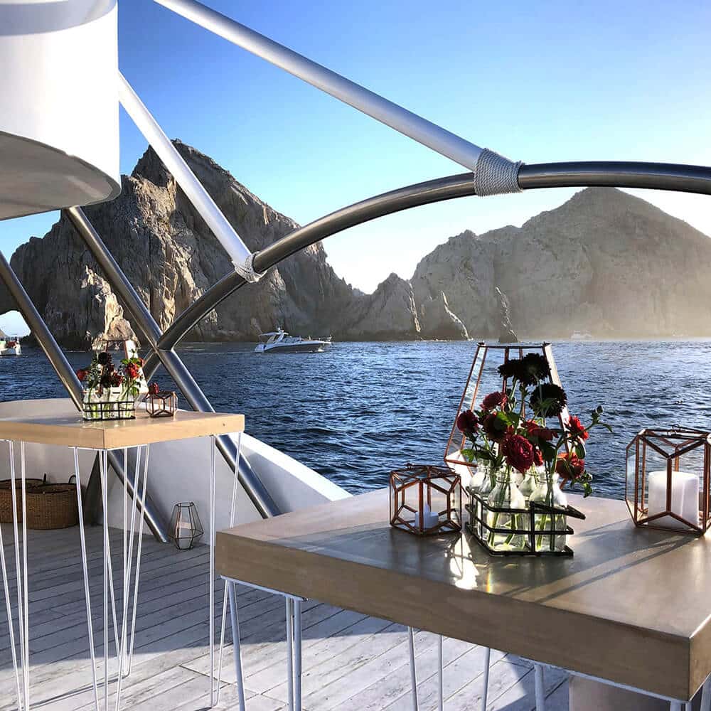 High tables with flower arrangements and lanterns on deck, rocks and a boat in the distance