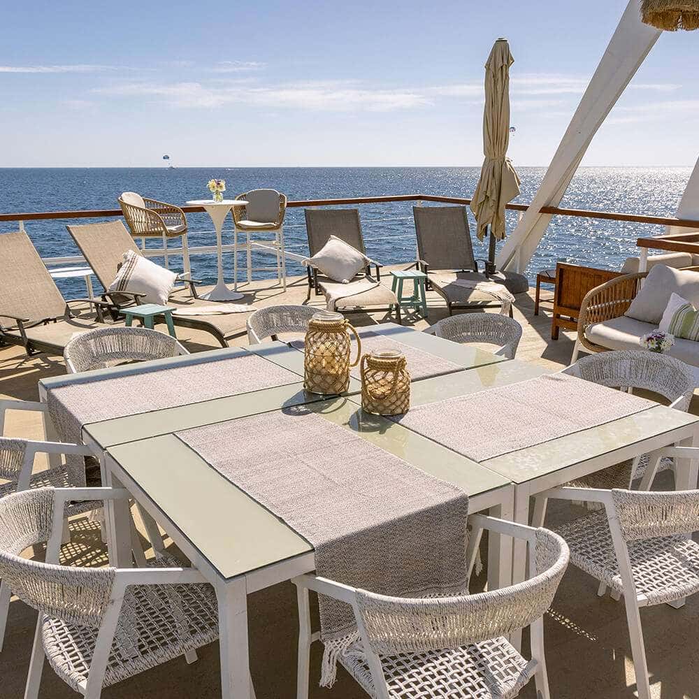 Long dining table with white chairs on deck, loungers and the sea behind