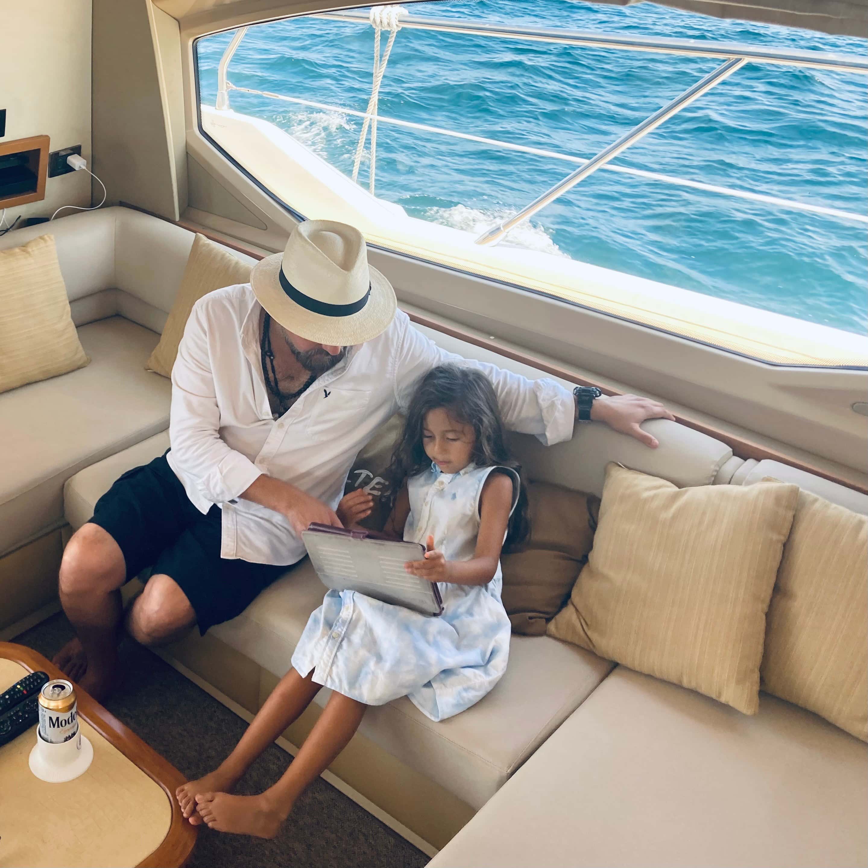 A man and a child on a cream sofa in the saloon, a window onto the sea beside them