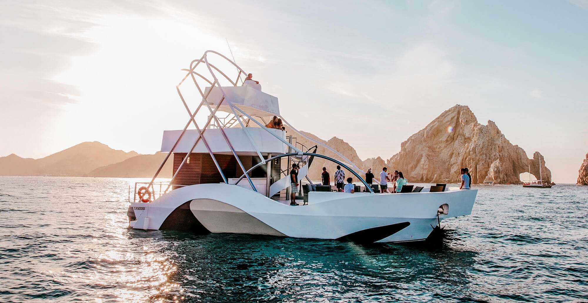 Pelican X catamaran at sunset with guests on deck, rocky Cabo San Lucas headland and arch behind