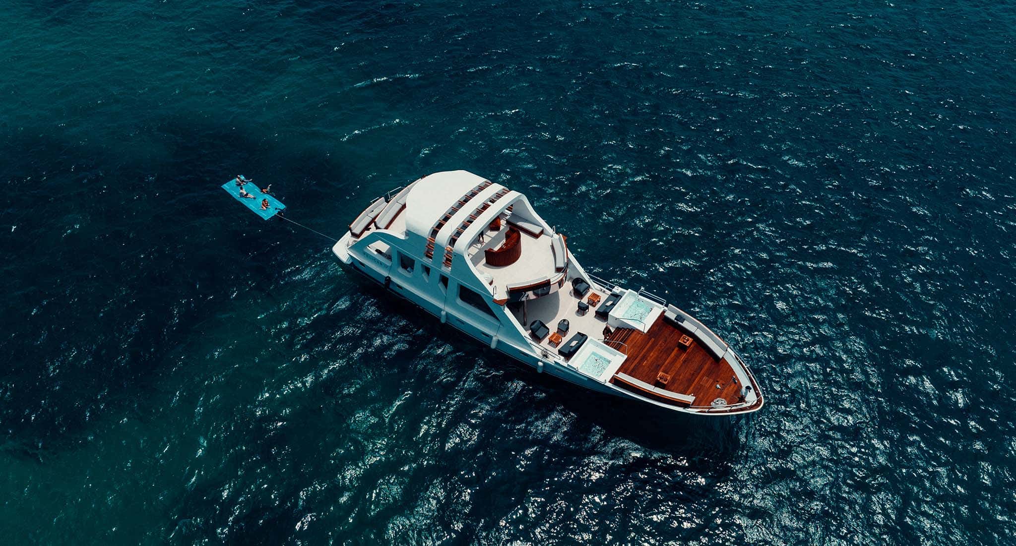 Aerial view of Pelican 17 on dark teal water, towing a blue floating mat with swimmers
